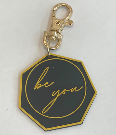 Keychain made out of gold and black PVC, with a gold clip and Be You written across the center.