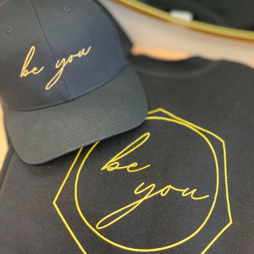 A black snapback hat with 'be you' embroidered in gold on the front and a black crewneck with the Be You logo screen printed on the chest in gold.